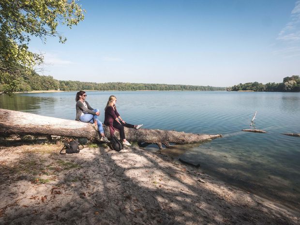 Zwei junge Frauen machen Pause auf einem Baumstamm am Ufer des Straussees. 